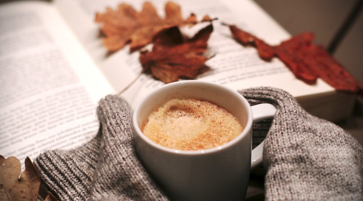 Tasse chaude entre des mains en pull de laine, livre ouvert et feuilles d’automne