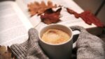 Tasse chaude entre des mains en pull de laine, livre ouvert et feuilles d’automne